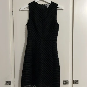 Svart ärmlös klänning från H&M - Svart kortklänning från H&M med snyggt strukturmönster i diagonala ränder. Klänningen är ärmlös, har rund halsringning och en guldig dragkedja i ryggen. Materialet är polyester med ett fodrat inre lager för extra komfort.
