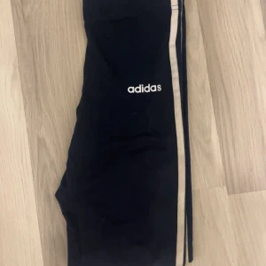 Svarta leggings från Adidas - Snygga svarta leggings från Adidas med klassiska vita ränder längs sidorna och Adidas-logga på låret. Perfekta för träning eller chill, med tight passform och stretchigt material som ger skön rörelsefrihet.