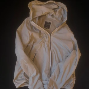 Ljusbeige vindjacka från Boggi Milano - Snygg ljusbeige vindjacka från Boggi Milano med huva och dragkedja framtill. Jackan har två sidofickor med dragkedja och meshfoder på insidan för extra komfort. Perfekt för dig som gillar stilren och sportig look.