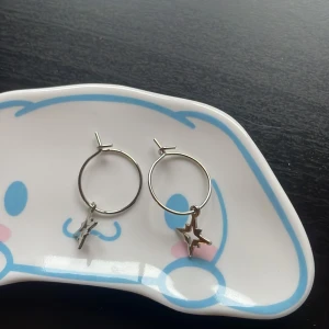 Silvriga örhängen med stjärnhänge - Ett par stilrena silvriga örhängen i ringmodell med hängande stjärnformad detalj. Aldrig användna har enbart haft på smyckestallrik, dock har ena en liten missfärgningsfläck (se bilaga 2) men som knappt syns när man har på dem! Frågor? Kom DM! 