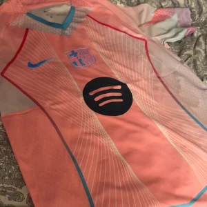 Barcelona Nike fotbollströja rosa - Snygg Barcelona fotbollströja från Nike i rosa med ljusblå och röda detaljer. Tröjan har klubbmärke, Nike-logga och Spotify som sponsor på bröstet. Tillverkad i lätt och ventilerande material, perfekt för match eller träning.