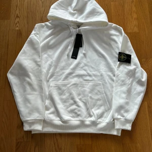 Vit Stone Island hoodie  - Säljer en clean vit hoodie från Stone Island med klassisk patch på vänster ärm. Hoodien har känguruficka, snörning i huvan och ribbade muddar. Perfekt för dig som gillar streetwear och vill ha en stilren look.