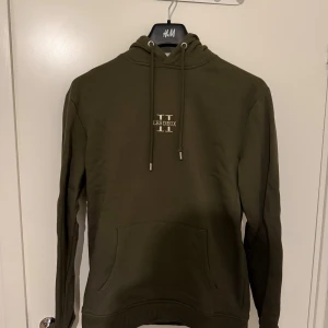 Olivgrön hoodie från Les Deux - Snygg olivgrön hoodie från Les Deux med broderad logga på bröstet. Tröjan har huva med snörning, känguruficka och långa ärmar. Perfekt för dig som gillar en clean och stilren look. Litet skavanker på högra snöret därav billigare pris