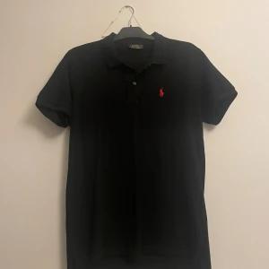 Svart pikétröja från Polo Ralph Lauren - Svart pikétröja från Polo Ralph Lauren med klassisk krage och två knappar framtill. Ikoniska röda broderade loggan på bröstet. Tillverkad i mjuk bomull och har korta ärmar. Perfekt för en clean och stilren look.