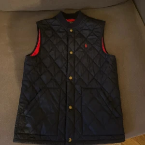 Quiltad svart väst från Polo Ralph Lauren - Snygg quiltad väst från Polo Ralph Lauren i svart med röd insida och guldfärgade knappar. Västen har två fickor fram och ett diskret rött Polo-logo broderat på bröstet. Perfekt för lager-på-lager och har en klassisk, stilren look.