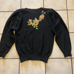 Svart stickad tröja med broderade blommor storlek S/M - Svart stickad tröja med lång ärm och rund hals. På framsidan finns broderade blommor i gult och orange med gröna blad, vilket ger en snygg och unik detalj. Tröjan har ribbade muddar vid ärmslut och nederkant. Perfekt för dig som gillar coola broderier och vill sticka ut.