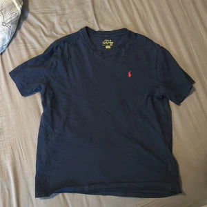 Mörkblå t-shirt Polo Ralph Lauren - Snygg mörkblå t-shirt från Polo Ralph Lauren med klassisk v-ringning och röd broderad logga på bröstet. Tillverkad i mjuk bomull och har en normal passform. Perfekt för en clean och stilren look.