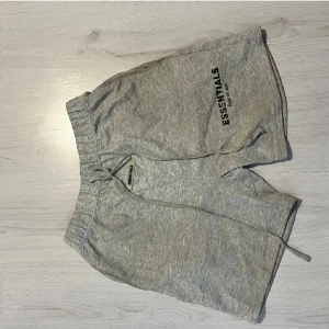 Grå shorts Essentials Fear of God - Snygga grå shorts från Essentials Fear of God med tryckt logga på ena benet. De har elastisk midja med snörning och är tillverkade i mjuk bomull. Perfekta för chill dagar eller när du vill ha en clean streetwear-look.