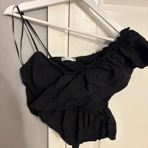 Svart oneshoulder croppad topp från Zara - Säljer en svart croppad topp från Zara med oneshoulder-design och tunna axelband. Toppen har volangdetaljer och en snygg rynkad effekt längs axeln och midjan. Perfekt för dig som gillar trendiga och unika plagg med cool siluett.