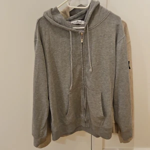 Grå hoodie - Grå hoodie i storlek XL med dragkedja framtill, snörning i huvan och klassisk logga på ärmen. Tröjan har känguruficka och är tillverkad i mjuk bomull. Perfekt för en chill och avslappnad stil. Passar även dem som har Ł.