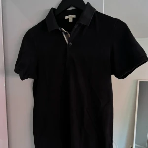 Burberry Piké - Klassisk svart pikétröja från Burberry med korta ärmar och ikonisk rutdetalj vid knäppningen. Säljer då den ej kommer till använding! hyfsat bra skick, inga flaws eller sånt. Pris kan diskuteras vid snabb affär!