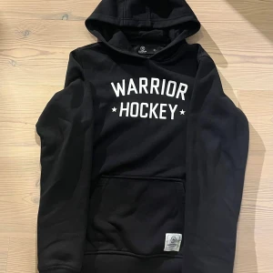 Svart hoodie Warrior Hockey 160 - Svart hoodie från Warrior Hockey i storlek XL motsvarar ungefär storlek 160.   Tröjan har huva, magficka och vit text framtill. Mjuk och skön. Perfekt till hockey.Klassisk passform med långa ärmar och ribbade muddar. Snygg logga på fickan.