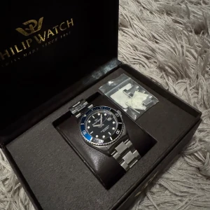 Philip Watch blå-silver klocka - Stilren klocka från Philip Watch med blå urtavla och silverfärgad boett och länkarmband i metall. Klockan har tydliga index, vridbar bezel i blått och silver samt vattentäthet upp till 100 meter. Köpt av Nilan Sthlm för 2500kr.Kommer i originalask med extra länkar.