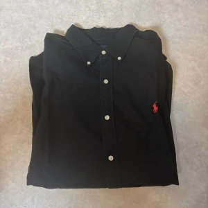 Svart skjorta från Ralph Lauren XL - Svart skjorta från Ralph Lauren i slim fit med klassisk krage och vita knappar. Broderad röd logga på bröstet ger en snygg kontrast. Skjortan är långärmad och tillverkad i mjuk bomull, perfekt för dig som gillar stilrena plagg med en ikonisk touch.