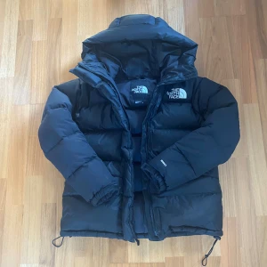 The North Face Vinterjacka  - Säljer en svart dunjacka från The North Face i storlek M. Jackan har en rejäl huva, dragkedja framtill och elastiska muddar vid ärmsluten. Klassisk pufferstil med logga på bröstet och ryggen. Perfekt för kalla dagar när du vill hålla värmen.