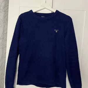 Marinblå stickad tröja från GANT - Snygg marinblå stickad tröja från GANT i storlek M. Tröjan har ett subtilt zigzag-mönster och rund halsringning. GANT-loggan är broderad på bröstet. Perfekt för dig som gillar klassisk stil med en twist.