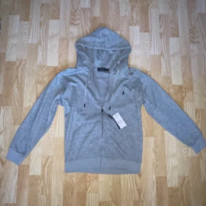 Grå hoodie från Polo Ralph Lauren - Snygg grå hoodie från Polo Ralph Lauren i storlek M. Tröjan har huva med snörning, dragkedja framtill och det klassiska Polo-logot broderat på bröstet. Tillverkad i mjuk bomull och har långa ärmar med ribbade muddar. Perfekt för chill dagar. Passformen är ganska liten så jag skulle rekommendera att köpa den om du har storlek S.