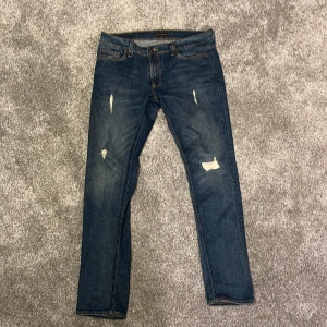 Nudie Jeans blå slitna jeansbyxor - Säljer ett par blå jeans från Nudie Jeans med snygga slitningar och detaljer på benen. Modellen har klassisk femficksdesign, orange sömmar och broderade bakfickor. Passformen är rak och materialet är robust denim. Perfekt för dig som gillar en cool och avslappnad stil.