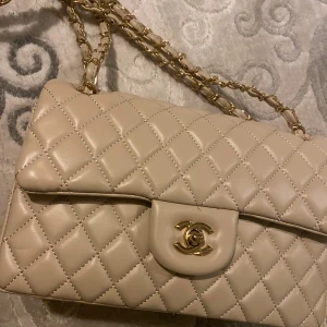 Väska - Supersnygg beige axelväska guld detaljer. Väskan har kedjerem med skinninslag och innerfack med dragkedja. Perfekt för dig som vill ha en ikonisk och lyxig accessoar med tidlös design. Öppen för prisförslag!