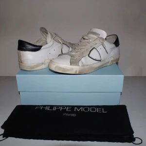 Philippe Model vita sneakers med svart detalj - Snygga vita sneakers från Philippe Model med svart häl och grå mockadetaljer. Skorna har rund tå, platt sula och klassisk snörning. Materialmix av skinn och mocka ger en cool och lyxig vibe. Perfekt för dig som gillar stilrena och trendiga sneakers.