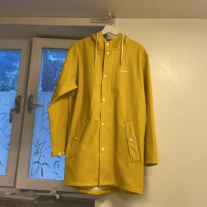 Gul regnjacka från Tretorn med huva - Snygg gul regnjacka från Tretorn med vit knappstängning, två fickor och justerbar huva med snören. Jackan är lång och har en clean look med raka linjer och vita detaljer. Perfekt för regniga dagar och håller dig torr med stil. Jackan har använts endast två gånger. Observera jackan har små fläckar i luvan som möjligt går att tvätta bort. 