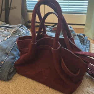 Vinröd mocka handväska med gulddetaljer - (Inte äkta) Snygg vinröd handväska i mjuk mocka med guldfärgade metalldetaljer. Väskan har två rejäla handtag och en axelrem, samt magnetlås och innerfack. Perfekt storlek för att få plats med allt du behöver och ger en lyxig vibe till din outfit.