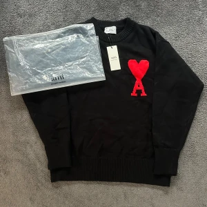 Svart stickad tröja från Ami - Svart stickad tröja från Ami Alexandre Mattiussi med ett stort rött hjärta och bokstaven A broderat på bröstet. Tröjan har rund hals, ribbade muddar och nederkant samt långärmad passform. Perfekt för dig som gillar stilrena detaljer med en twist.