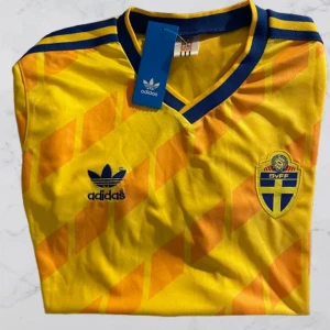 Sverige fotbollströja retro - Gul fotbollströja från Adidas med blå detaljer och SVFF-emblem på bröstet. Tröjan har korta ärmar, V-ringad krage med blå kant och ett snyggt diagonalt mönster i orange. Materialet är lätt polyester, perfekt för match eller träning. NYTT SKICK!