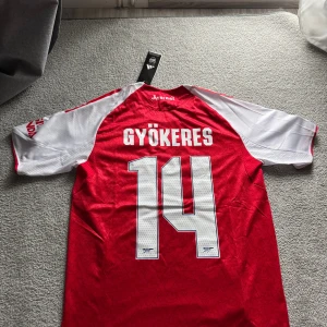 Arsenal Hemmaställ 25/26 Gyökeres Champions League - Röd och vit Arsenal fotbollströja från Adidas med Gyökeres och nummer 14 på ryggen. Tröjan har korta ärmar, broderat klubbmärke, Champions League-märke på ärmen och klassiska Adidas-ränder. Tillverkad i lätt och ventilerande polyester.