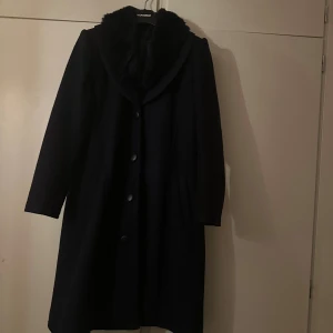 Marinblå kappa med fuskpäls  - Stilren svart kappa med bred krage i mjuk fuskpäls. Kappan har knäppning framtill med stora knappar och en klassisk, något figursydd passform. Perfekt för kalla dagar när du vill vara både varm och snygg.