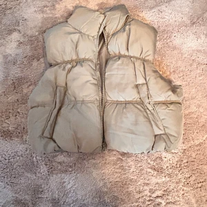Beige dunväst från H&M Divided - Säljer en beige dunväst från H&M Divided i storlek S. Västen har en hög krage, dragkedja framtill och två fickor på sidorna. Perfekt att slänga över en hoodie för en cool och avslappnad look.