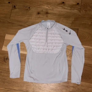 Nike tröja - Ljusgrå långärmad träningströja från Nike med Tottenham Hotspur-logga på ärmen. Tröjan har quiltad frontpanel, half zip och blå detaljer längs ärmarna. Perfekt för fotbollsträning eller chill dagar. Materialet är polyester och syntet.