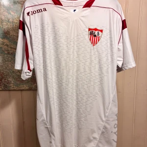 Sevilla FC 2007/08 Joma home shirt, size 3XL - Säljer en vit Sevilla FC fotbollströja från Joma i storlek 3XL. Tröjan har röda detaljer på axlar och ärmar, klubbmärke på bröstet och diskret textmönster över hela framsidan. Tillverkad i lätt och ventilerande polyester, perfekt för match eller träning.