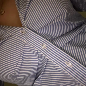 Blårandig skjorta med knappar - Snygg skjorta med smala blå och vita ränder, klassisk krage och knappar framtill. Skjortan har lång ärm och figurnära passform som framhäver midjan. Perfekt för dig som gillar stilrena och fräscha plagg med lite edge.