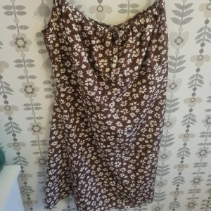 Brun blommig klänning med snörning - Säljer en brun klänning med vita blommor och snörning vid halsen. Klänningen har en avslappnad passform och är ärmlös, perfekt för varma dagar. Materialet känns lätt och luftigt, och mönstret ger en skön retrovibe.
