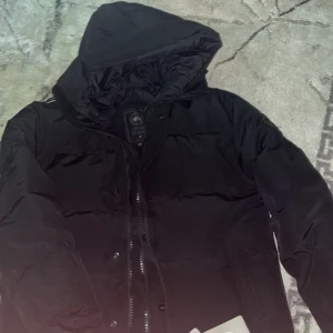 Svart dunjacka från Canada Goose - Svart dunjacka från Canada Goose med huva och klassisk logotyp på ärmen. Jackan har dragkedja och knappar framtill, samt två sidofickor. Perfekt för kalla dagar med sin tjocka och puffiga design. Snygg och stilren look som funkar till det mesta.