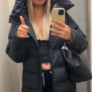 Svart lång pufferjacka med huva - Säljer en svart lång pufferjacka med stor huva och dragkedja framtill. Jackan har quiltad design, slits med knappar på sidan och är perfekt för kalla dagar. Den har en loose passform och är tillverkad i syntetmaterial. Grym att bära över hoodies eller stickade tröjor.