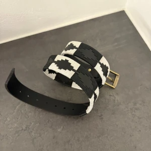 Svart och vit mönstrad skinnbälte från Pampeano - Säljer ett svart skärp i skinn med coolt geometriskt mönster i vitt. Bältet har en robust mässingsfärgad metallspänne och detaljerad brodyr längs hela längden. Perfekt för att ge outfiten en unik touch.