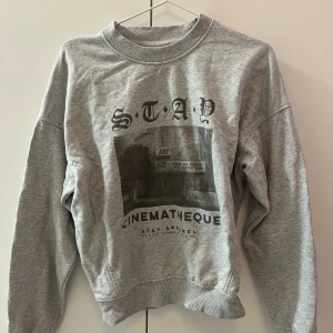 Grå sweatshirt från Stay  - Snygg grå sweatshirt från Stay med tryck på framsidan som föreställer en biograf och texten 'Cinematheque'. Tröjan har rund hals, ribbade muddar och lång ärm. 