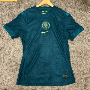 Nigeria Nike fotbollströja blågrön - Säljer en officiell helt ny Nigeria Football Federation fotbollströja från Nike i blågrön färg och med etikett. Tröjan har korta ärmar, subtilt mönster i tyget och limegröna detaljer som logga och emblem. Tillverkad i lätt och ventilerande Dri-FIT-material, perfekt för träning eller match. Skriv vid minsta fundering. Priset kan diskuteras!
