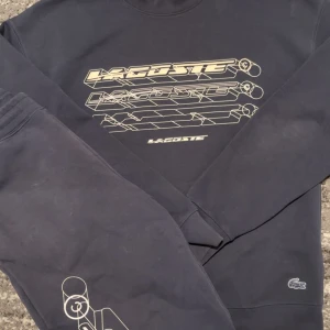 Svart loose fit sweatshirt från Lacoste - Cool svart sweatshirt från Lacoste med loose fit och grafiskt tryck i vitt på bröstet. Tröjan har rund hals, långa ärmar och den klassiska Lacoste-loggan broderad nertill. Perfekt för en avslappnad streetstyle-look.