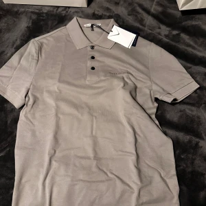 Grå pikétröja från Tiger of Sweden - Snygg grå pikétröja från Tiger of Sweden med klassisk krage och tre knappar framtill. Tröjan har korta ärmar och diskret logga på bröstet. Perfekt för en clean och stilren look.