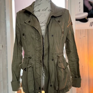 Olivgrön parkajacka från Hollister - Säljer en olivgrön parkajacka från Hollister med dragsko i midjan, stora fickor och snygga detaljer vid axlar och ärmslut. Jackan har dragkedja och knappar framtill samt ett diskret Hollister-märke på ärmen. Perfekt för vår och höst!