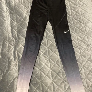 Nike Pro Hyperwarm leggings svart/grå S - Nike Pro Hyperwarm leggings i storlek S med snygg ombre-effekt från svart upptill till ljusgrå nedtill. Stretchigt och värmande material, perfekt för träning. Nike-logga på benet och bred resår med Nike Pro-tryck i midjan.