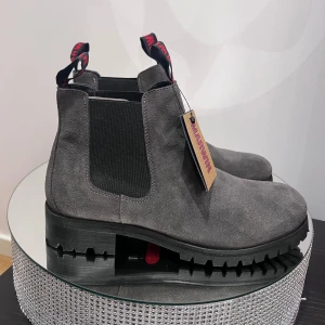 Grå Chelsea boots från Marwin - Snygga Chelsea boots i mörkgrå mocka från Marwin med kraftig platt sula och elastiska paneler på sidorna. Skorna har rund tå och tydliga dragflikar med röd logga baktill för enkel påtagning. Perfekta för dig som gillar stilren och robust design.