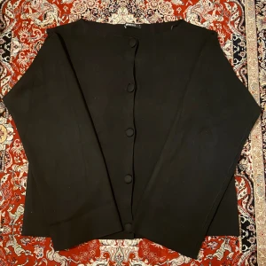 Svart kofta med knappar från Gina Tricot - Stilren svart kofta från Gina Tricot i storlek S. Koftan har långa ärmar, rund halsringning och knäppning framtill med tygklädda knappar. Perfekt att slänga över en topp för en clean look.