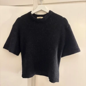 Svart stickad tröja från Gina Tricot - Mysig svart stickad tröja från Gina Tricot med korta ärmar och rund hals. Tröjan har en enkel och stilren design, materialet känns mjukt och fluffigt, vilket gör den extra skön att bära.