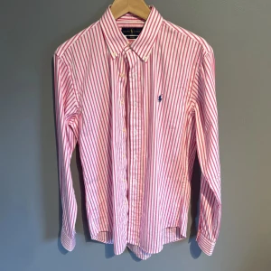 Rosa och vit randig skjorta Ralph Lauren - Snygg skjorta från Ralph Lauren i rosa och vitt med klassiska ränder. Skjortan har lång ärm, knappar framtill och den ikoniska blå loggan broderad på bröstet. Perfekt för dig som gillar en preppy och stilren look.