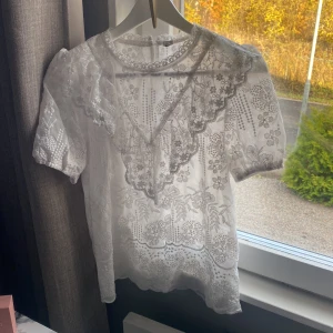 Vit blommig spetsblus - Superfin vit blus med blommigt Blusen har  spetsdetalj framtill och knäppning i nacken. 