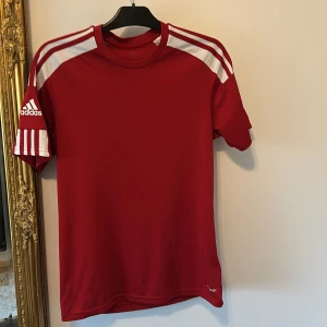 Röd Adidas fotbollströja med vita ränder - Röd fotbollströja från Adidas med klassiska vita ränder på axlarna och ärmarna. Tröjan har rund halsringning och är tillverkad i ett lätt och ventilerande funktionsmaterial. Adidas-logga på ena ärmen ger en sportig look.